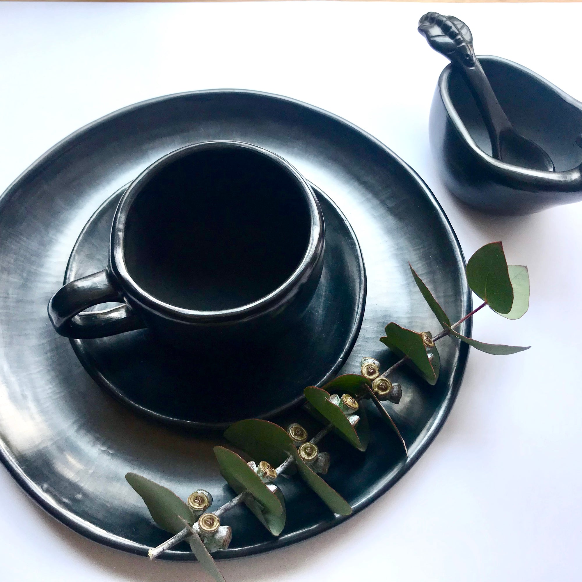 Oaxacan black clay plates, coffee cup and a salsa bowl with a rooster head, accompanied by a twig of fresh, light green-bluish eucalyptus 