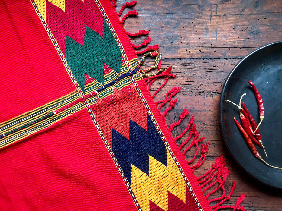Mexico 1492 - bright red table cloth with colorful edges, handmade on a backstrap loom in Chiapas Mexico. Paired with Black Clay plate from the same shop